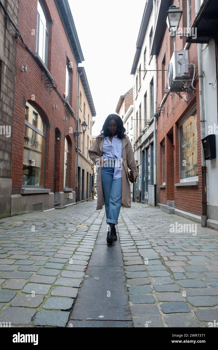 Dieses Bild zeigt eine stilvolle Frau, die eine Kopfsteinpflasterstraße in einem historischen europäischen Viertel entlang geht. Sie trägt ein Hemd mit blauem Kragen, Jeans mit hohem Bund, einen beigefarbenen Trenchcoat und schwarze Stiefel. Ihr selbstbewusster Schritt und die Perspektive des Fotos deuten auf Bewegung und ein Sinn für einen Zweck hin. Die traditionellen Gebäude mit Backsteinfassaden und Straßenlaternen verstärken das Ambiente der alten Welt. Stilvolle Frau, die in der historischen European Street läuft. Hochwertige Fotos Stockfoto
