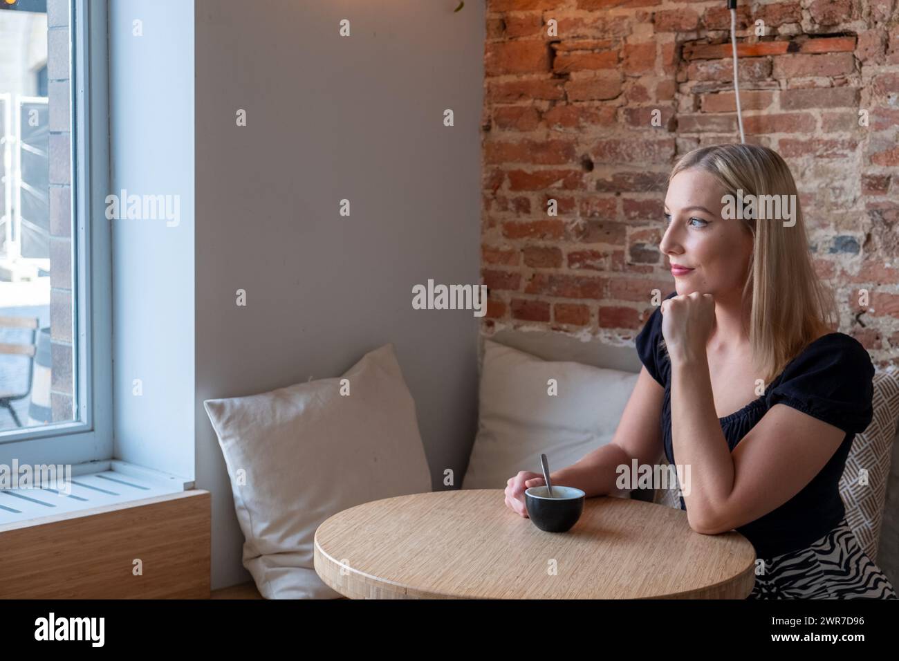 In einem ruhigen Café wird eine junge blonde Frau dargestellt, die einen Moment der Einsamkeit genießt. Sie sitzt an einem Holztisch und blickt nachdenklich aus einem großen Fenster, mit sanftem Tageslicht, das ihr Gesicht beleuchtet. Eine Tasse Kaffee wird sanft in ihren Händen gehalten, was auf eine friedliche Pause in ihrem Tag hindeutet. Die freiliegende Ziegelwand an ihrer Seite verleiht dem modernen Interieur einen rustikalen Charme und schafft eine gemütliche und einladende Atmosphäre. Die Einfachheit der Szene wird durch den Kontrast zwischen dem Blick auf die Stadt draußen und dem warmen, intimen Raum im Inneren bereichert. Besinnliche junge Frau genießt Kaffee in einem schicken Café. Hohe Qualität Stockfoto