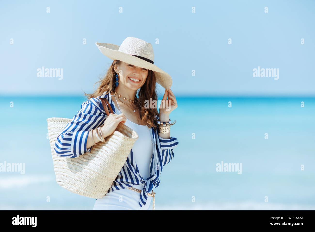 Lächelnde elegante 40-jährige Frau am Meer mit Strohsack und Strohhut. Stockfoto