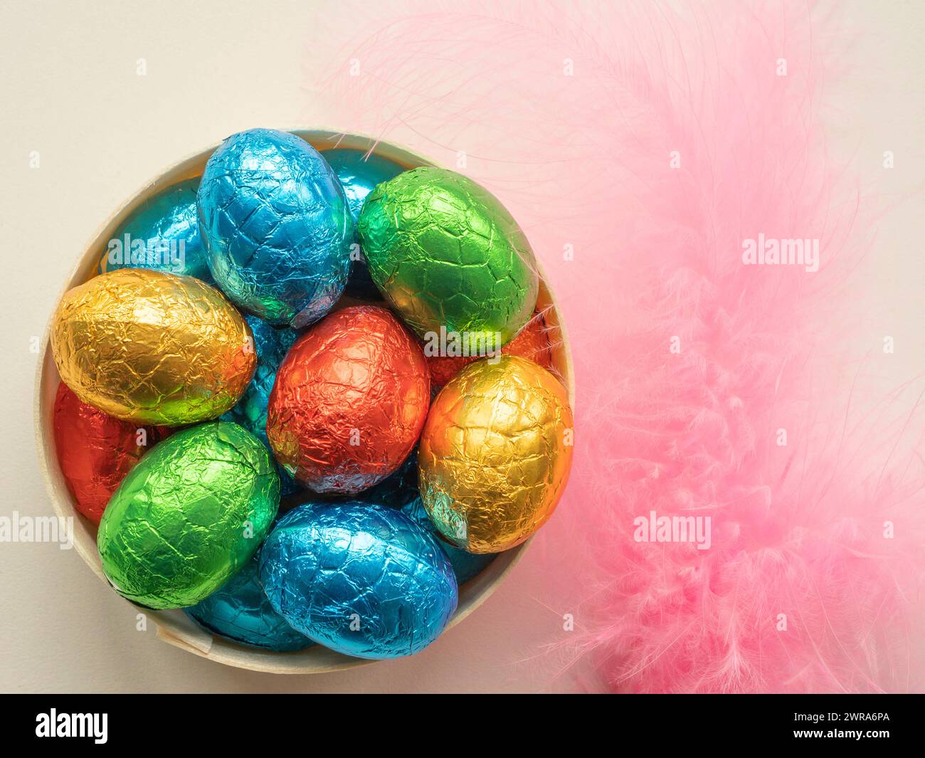 Hölzerne Schüssel gefüllt mit folienüberzogener Schokolade Ostereier mit rosafarbenen Federn Stockfoto