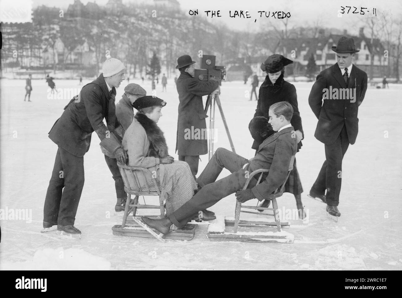 Auf dem Tuxedo-See sehen Sie Menschen, die auf Stühlen auf einem gefrorenen Teich mit Skatern im Tuxedo Park im Bundesstaat New York sitzen. Ein Mann mit einer Filmkamera filmt die Szene. 1910 und ca. 1920, Glasnegative, 1 negativ: Glas Stockfoto