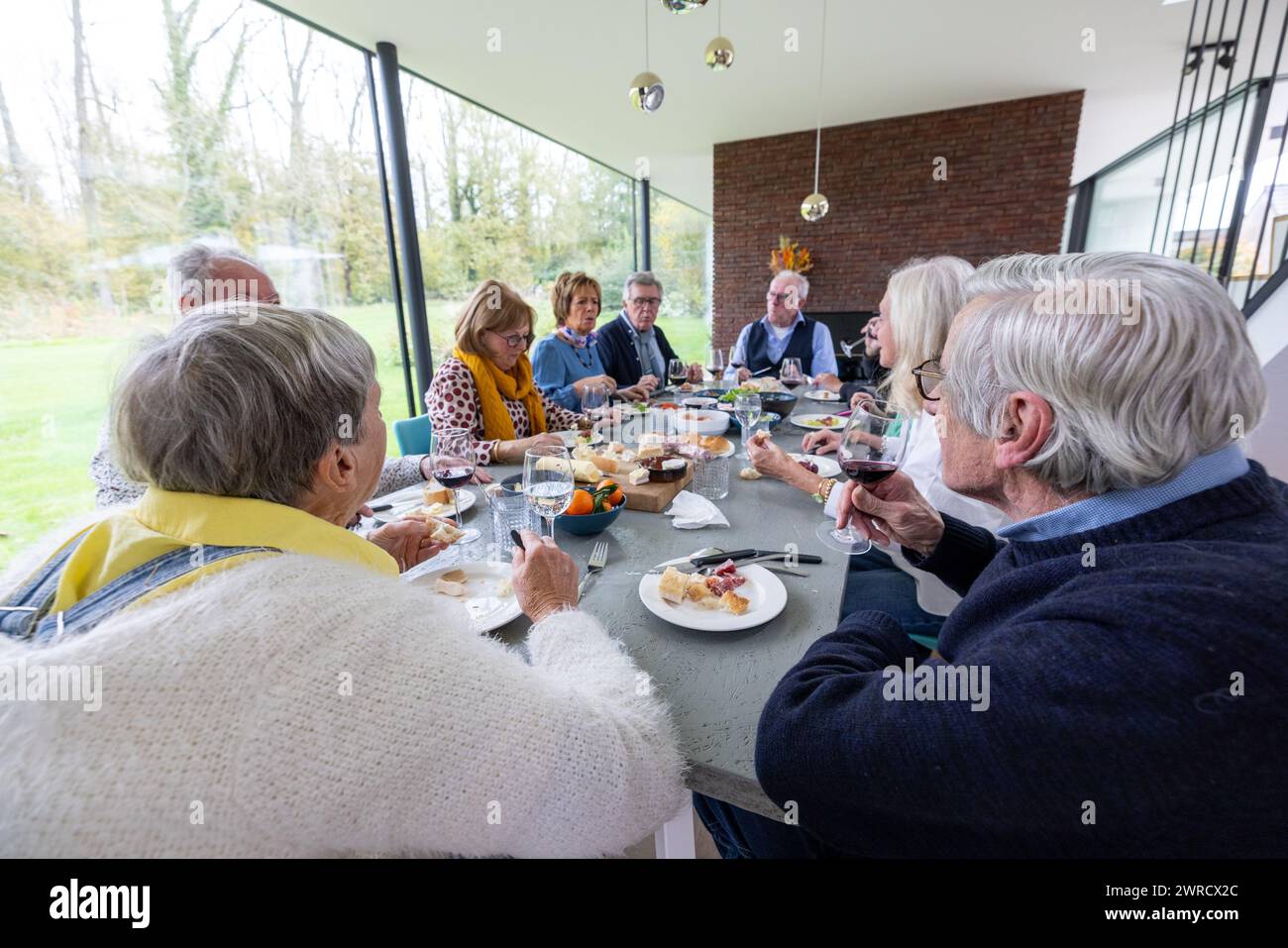 Gruppe von Erwachsenen, die sich an einem Esstisch unterhalten und gemeinsam eine Mahlzeit in einem modernen, gut beleuchteten Raum mit Blick auf die Natur genießen. Familientreffen für eine Mahlzeit in einem modernen, sonnendurchfluteten Raum. Hochwertige Fotos Stockfoto