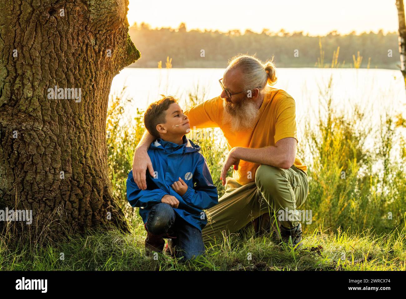 Dieses Bild fängt einen zärtlichen Moment zwischen einem Großvater und seinem Enkel ein, der bei Sonnenuntergang an einem ruhigen Seeufer sitzt. Der ältere Mann, mit weißem Bart, Brille und einem warmen Lächeln, legt seine Hand sanft auf die Schulter des Jungen und vermittelt ein Gefühl von Führung und Zuneigung. Der kleine Junge sieht ihn mit einem Blick der Bewunderung und des Vertrauens an. Sie sind von einem großen Baum eingerahmt, und die sanfte Hintergrundbeleuchtung der untergehenden Sonne schafft eine ruhige und warme Atmosphäre, die diesen intimen Familienmoment unterstreicht. Zärtlicher Moment zwischen Großvater und Enkel am See. Hochwertige Fotos Stockfoto