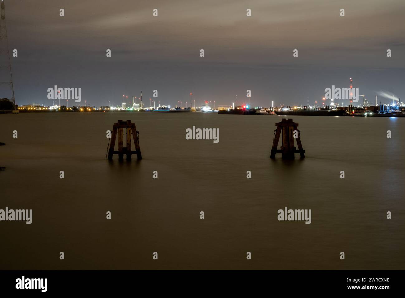 In diesem eindrucksvollen nächtlichen Bild sehen Sie einen Panoramablick auf einen geschäftigen Hafen über ruhiges Wasser. Industrielle Lichter funkeln in der Ferne und erzeugen einen lebendigen Kontrast zum dunklen Abendhimmel. Im Wasser steht ein Paar maritimer Strukturen, teilweise silhouettenförmig und geisterhaft, die als stiller Beobachter der unaufhörlichen Energie des Hafenbetriebs dienen. Die lange Exposition macht das Wasser weich und betont die Stille der Bucht im Vergleich zum fernen Treiben des Hafens. Harbor Nightscape: Industrial Glow Over Calm Waters. Hochwertige Fotos Stockfoto