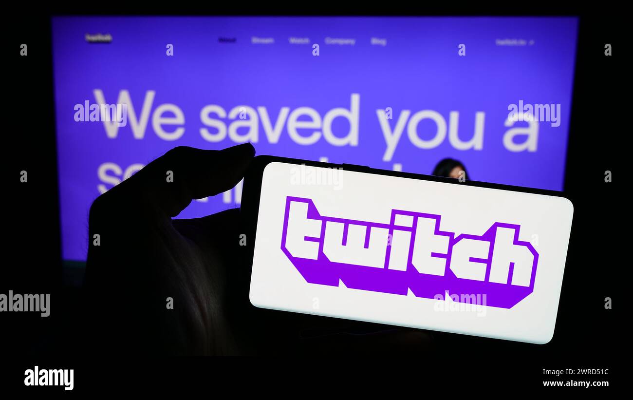 Person mit Mobiltelefon mit Logo des amerikanischen Live-Streaming-Unternehmens Twitch Interactive Inc. Vor der Webseite. Schwerpunkt auf dem Display des Telefons. Stockfoto