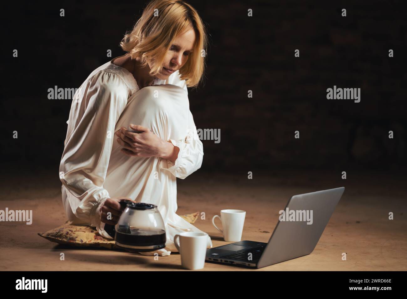 Eine Frau gießt Kaffee ein, während sie vor einem Laptop sitzt. Leerer Raum, dunkler Hintergrund. Deadline, Recycling, Fleiß-Freelancer-Konzept. Stockfoto