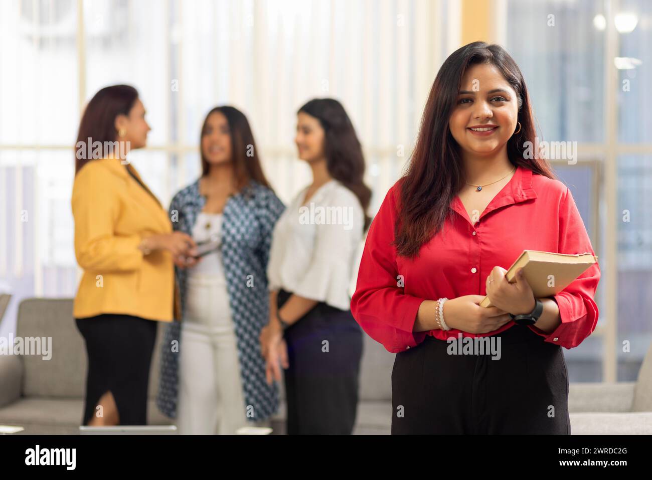 Porträt einer jungen Geschäftsfrau, die mit Notizbuch in einem Büro steht und ihre Kollegen im Hintergrund Stockfoto