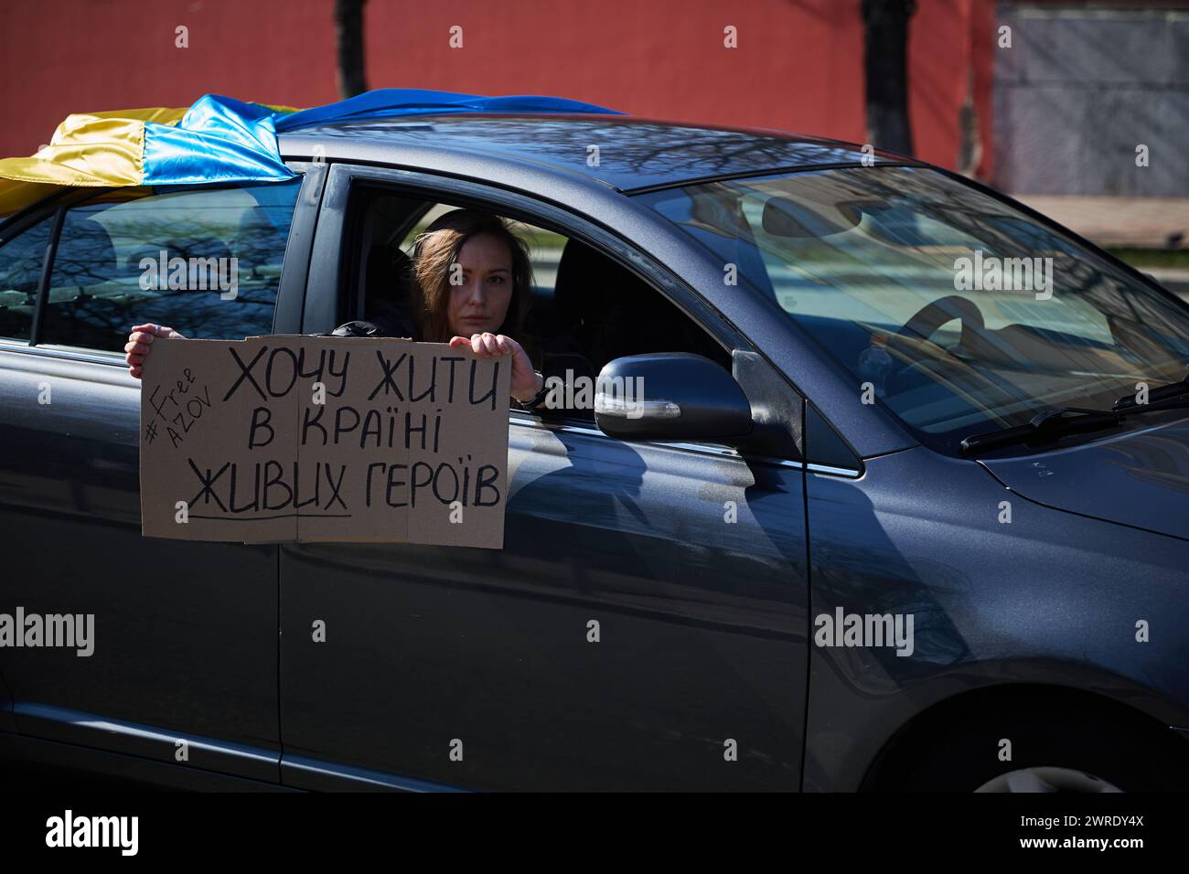 Die ukrainische Aktivistin zeigt auf einer öffentlichen Demonstration das Banner „Ich möchte in Einem Land lebendiger Helden leben“ aus einem Fenster eines fahrenden Fahrzeugs. Kiew - 10. März 2024 Stockfoto