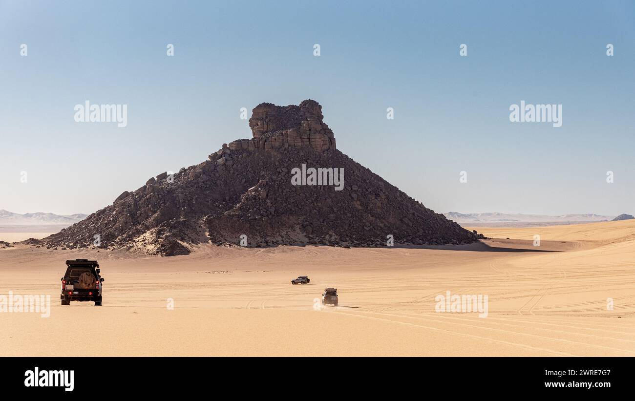 Landschaft der Region Tiboukaine in der Sahara, Algerien. Die Jeeps fahren auf einen Berg aus Geröll zu, der aus dem Sand zu entspringen scheint. Stockfoto