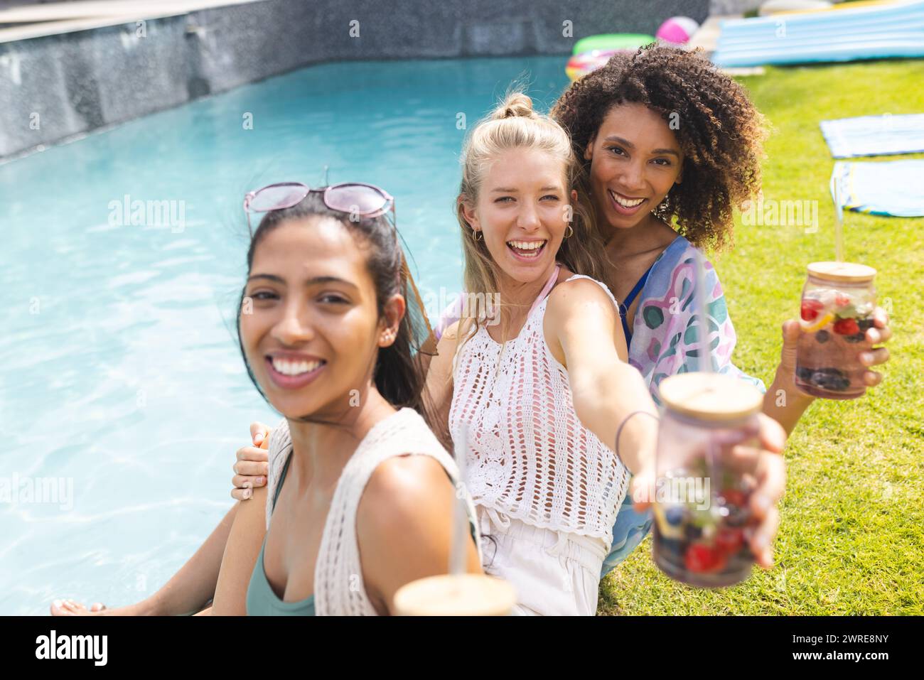 Verschiedene Freundinnen genießen ein sonniges Treffen am Pool und halten erfrischende Getränke Stockfoto