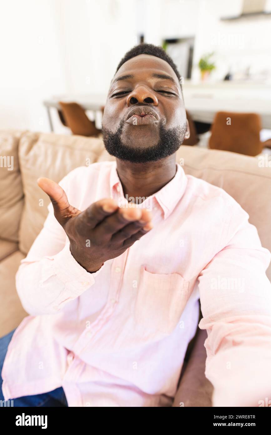Afroamerikaner in einem rosa Hemd spritzt einen Kuss auf einem Videoanruf, sitzt auf einem beigen Sofa Stockfoto