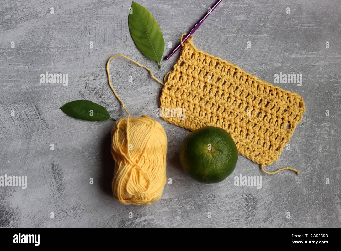 Häkelschal im Prozess. Häkelnadel, gelbes Baumwollgarn, Limette und grüne Blätter auf grauem Hintergrund, Draufsicht Stockfoto