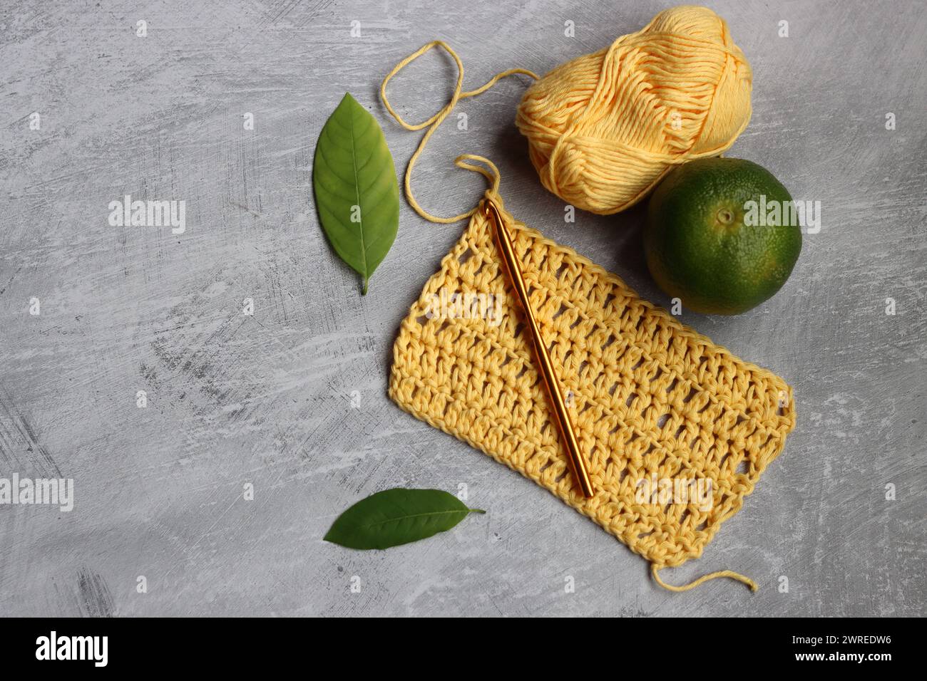 Häkelschal im Prozess. Häkelnadel, gelbes Baumwollgarn, Limette und grüne Blätter auf grauem Hintergrund, Draufsicht Stockfoto