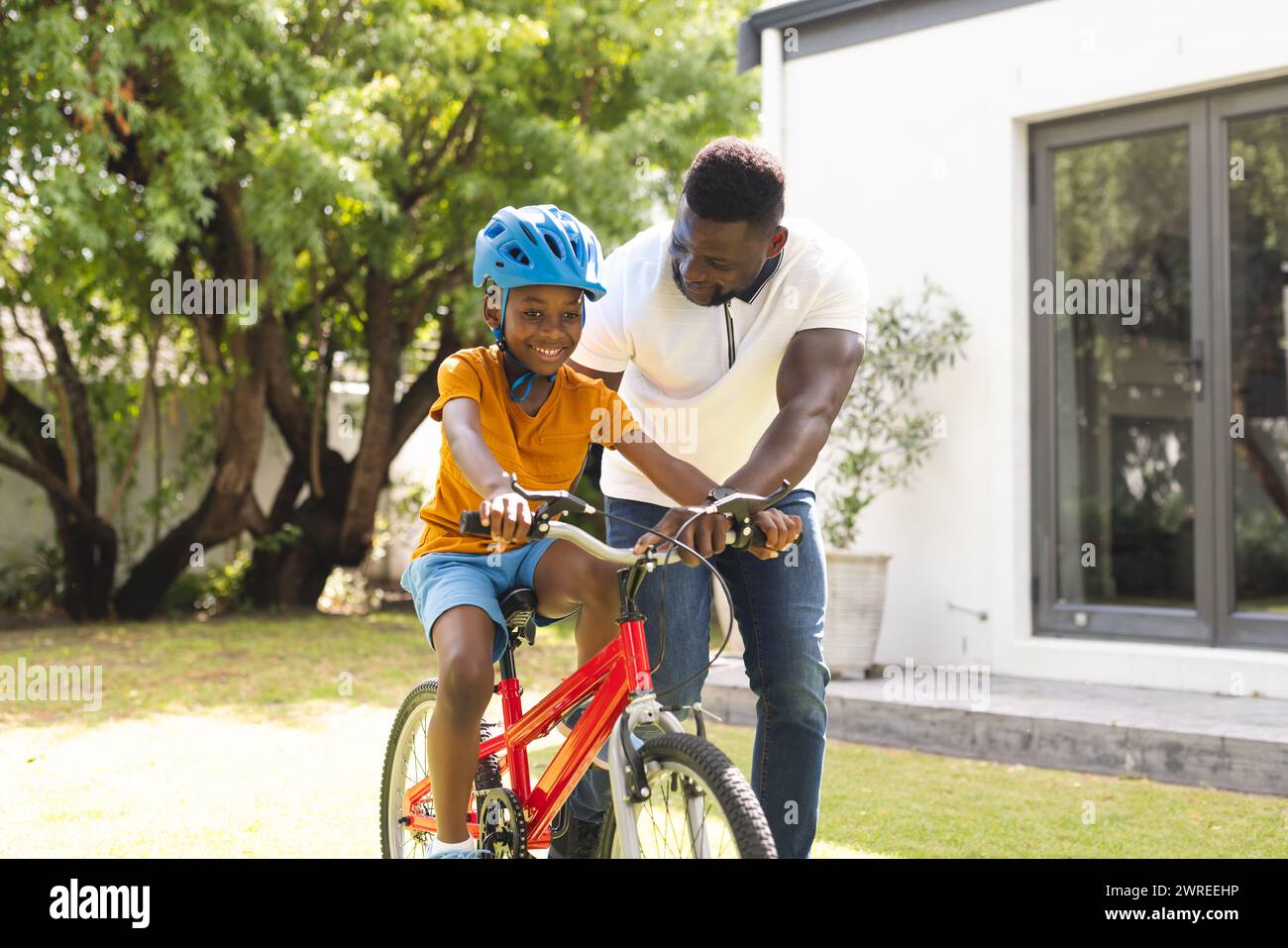Der afroamerikanische Vater hilft seinem Sohn, in einem sonnigen Hinterhof Fahrrad zu fahren Stockfoto