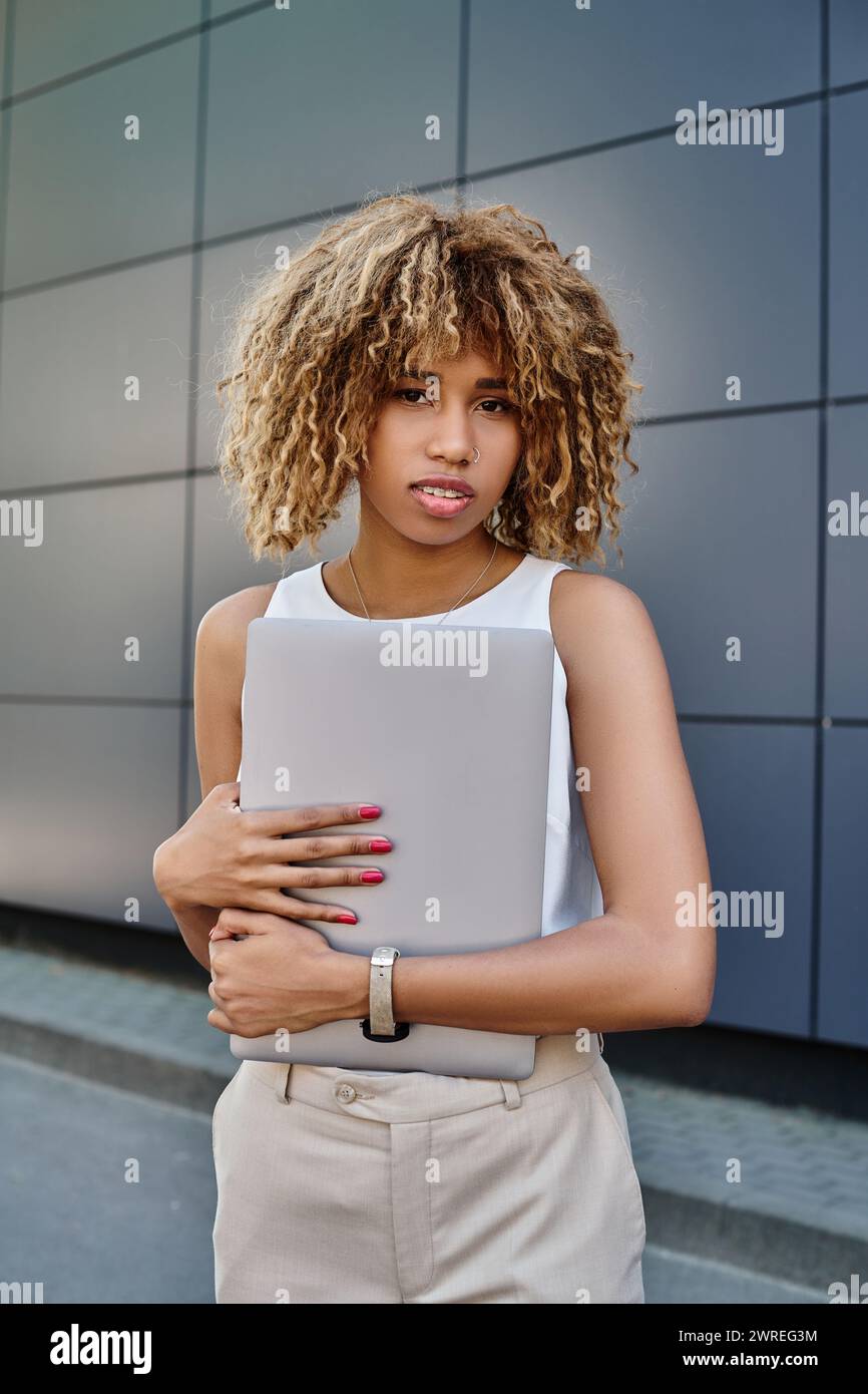 Lockige afroamerikanische Geschäftsfrau mit Zahnspange, die mit ihrem Laptop in der Nähe des Bürogebäudes steht Stockfoto