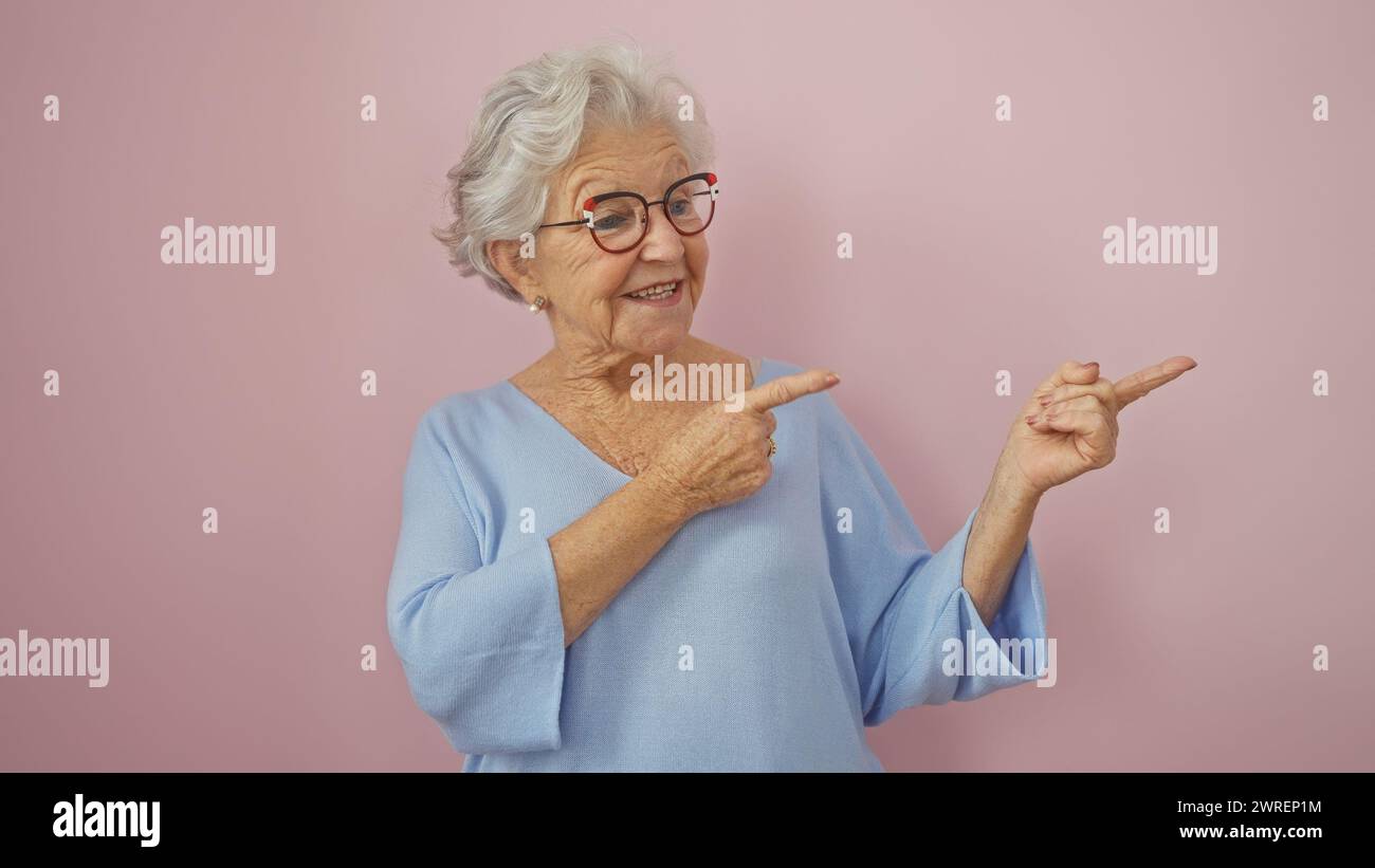 Eine fröhliche Seniorin in einem blauen Pullover, die glücklich auf einen rosa isolierten Hintergrund zeigt. Stockfoto