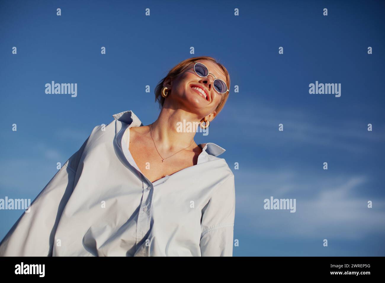 Moderne Frau in übergroßem Hemd und Sonnenbrille, fröhlich lächelnd am blauen Himmel. Trendige, fröhliche und stilvolle Sommeratmosphäre. Stockfoto