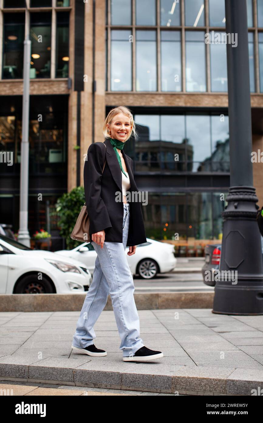 Wunderschöne junge, stilvolle Frau, die vor dem Hintergrund des Gebäudes die Straße entlang läuft, modische Kleiderjeans, schwarzen Blazer, Handtasche und Schnauze trägt Stockfoto