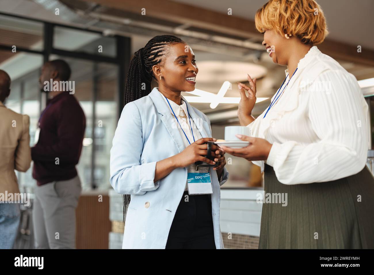 Zwei professionelle afrikanische Geschäftsfrauen führen ein Gespräch auf einer Geschäftskonferenz. Sie halten Kaffeetassen und tragen Konferenzabzeichen, Smili Stockfoto
