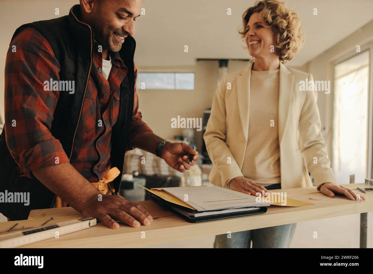 Ein männlicher Bauunternehmer und eine Hausbesitzerin arbeiten drinnen zusammen, lächeln und unterschreiben Dokumente. Die glücklichen Profis arbeiten gemeinsam an einer Hausrenovierung Stockfoto