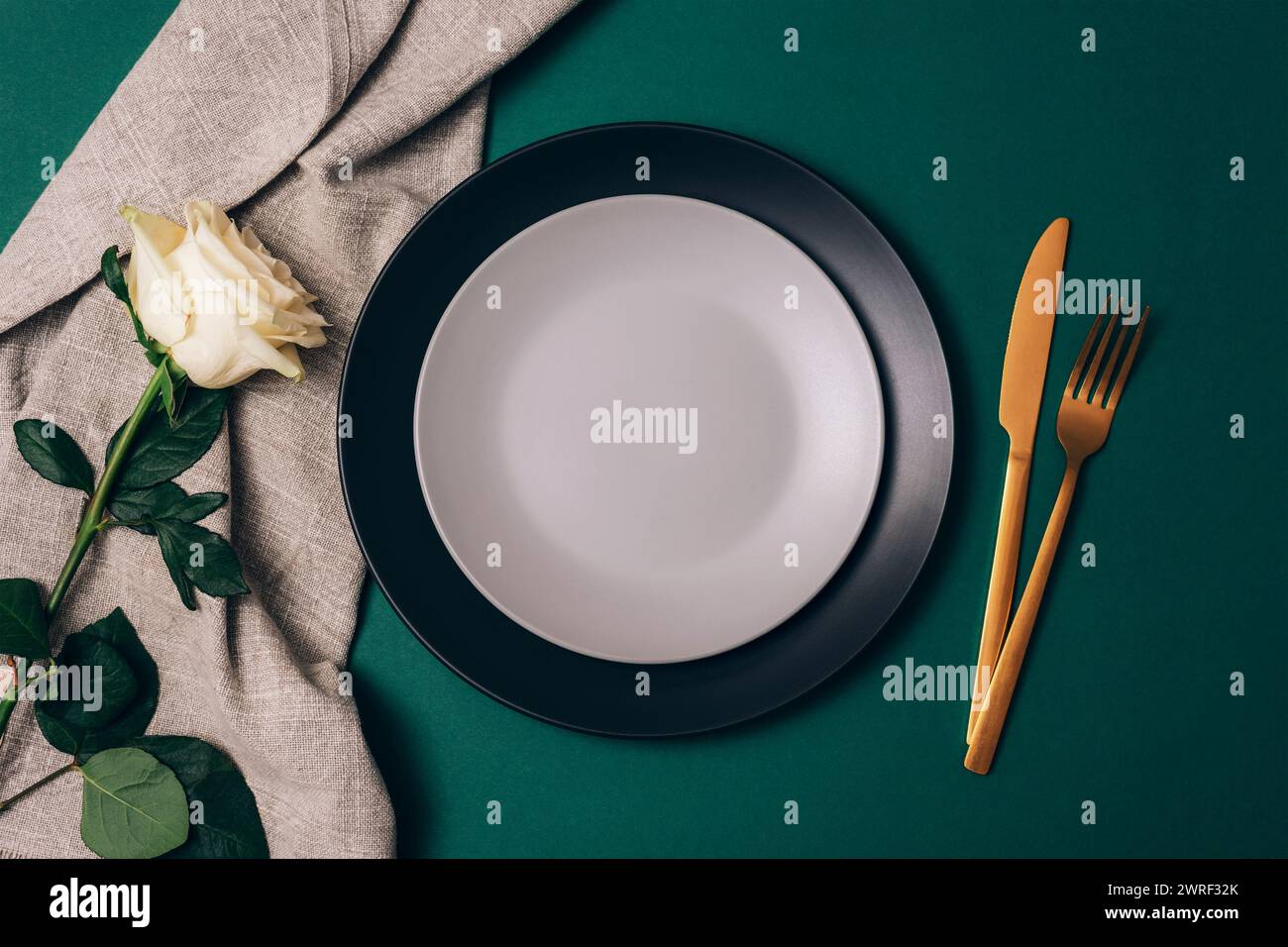 Tischdekoration mit weißer Rosenblüte, Tellern und goldenem Besteck auf dunkelgrünem Hintergrund. Draufsicht, flach. Stockfoto