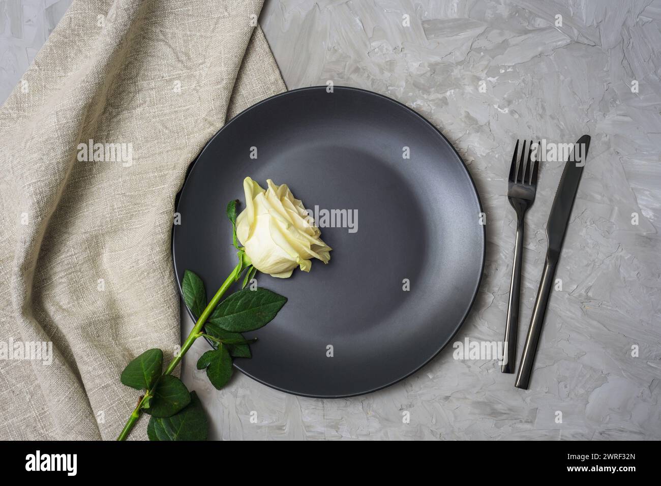 Tischdekoration mit weißer Rose, schwarzem Teller und Besteck auf strukturiertem grauen Hintergrund. Draufsicht, flach. Stockfoto