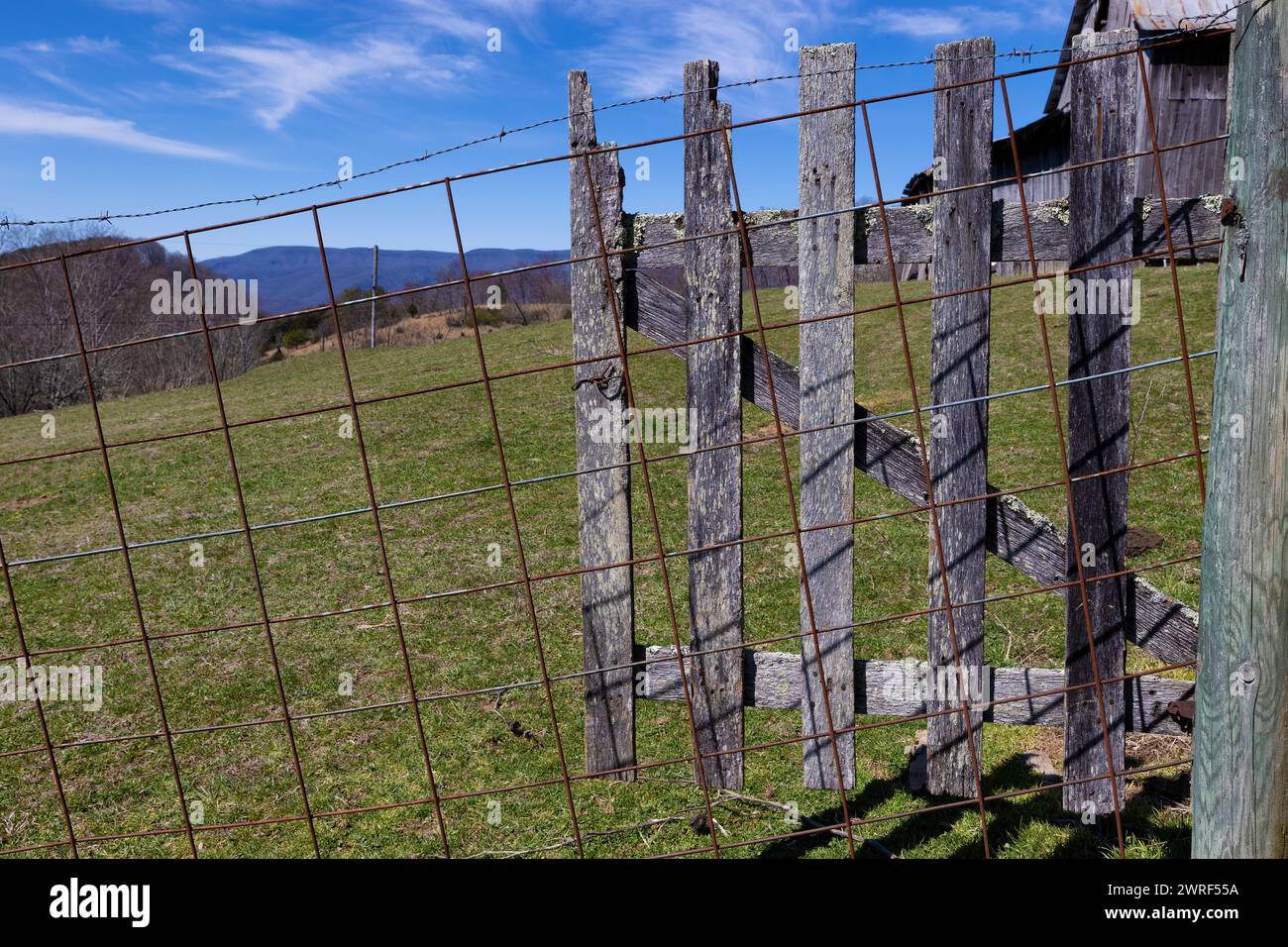 Der Blick auf die Landschaft und der Abschnitt einer alten Scheune kann durch den Drahtzaun und das offene Holztor im ländlichen Virginia gesehen werden. Stockfoto