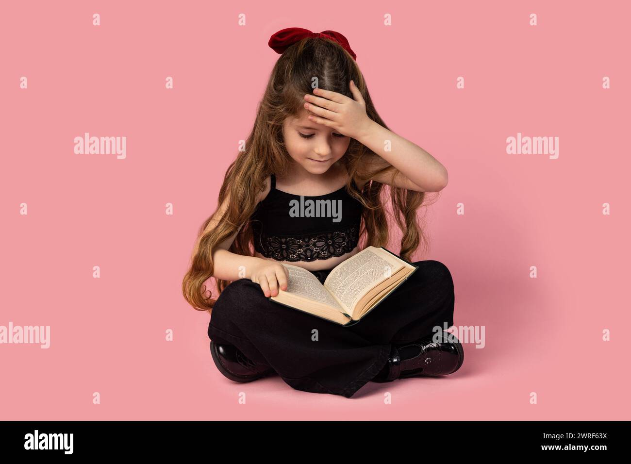 Das staunende Mädchen sitzt mit überkreuzten Beinen und der Hand auf der Stirn, schwarz gekleidet, studiert ein Buch, isoliert auf rosa Hintergrund. Hoch qua Stockfoto