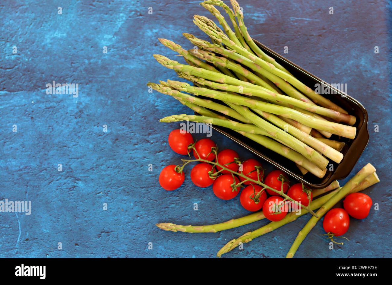 Frischer grüner Spargel und Kirschtomaten auf blau strukturiertem Hintergrund mit Kopierraum. Gesunde Zubereitung des Abendessens. Ausgewogenes Ernährungskonzept. Stockfoto
