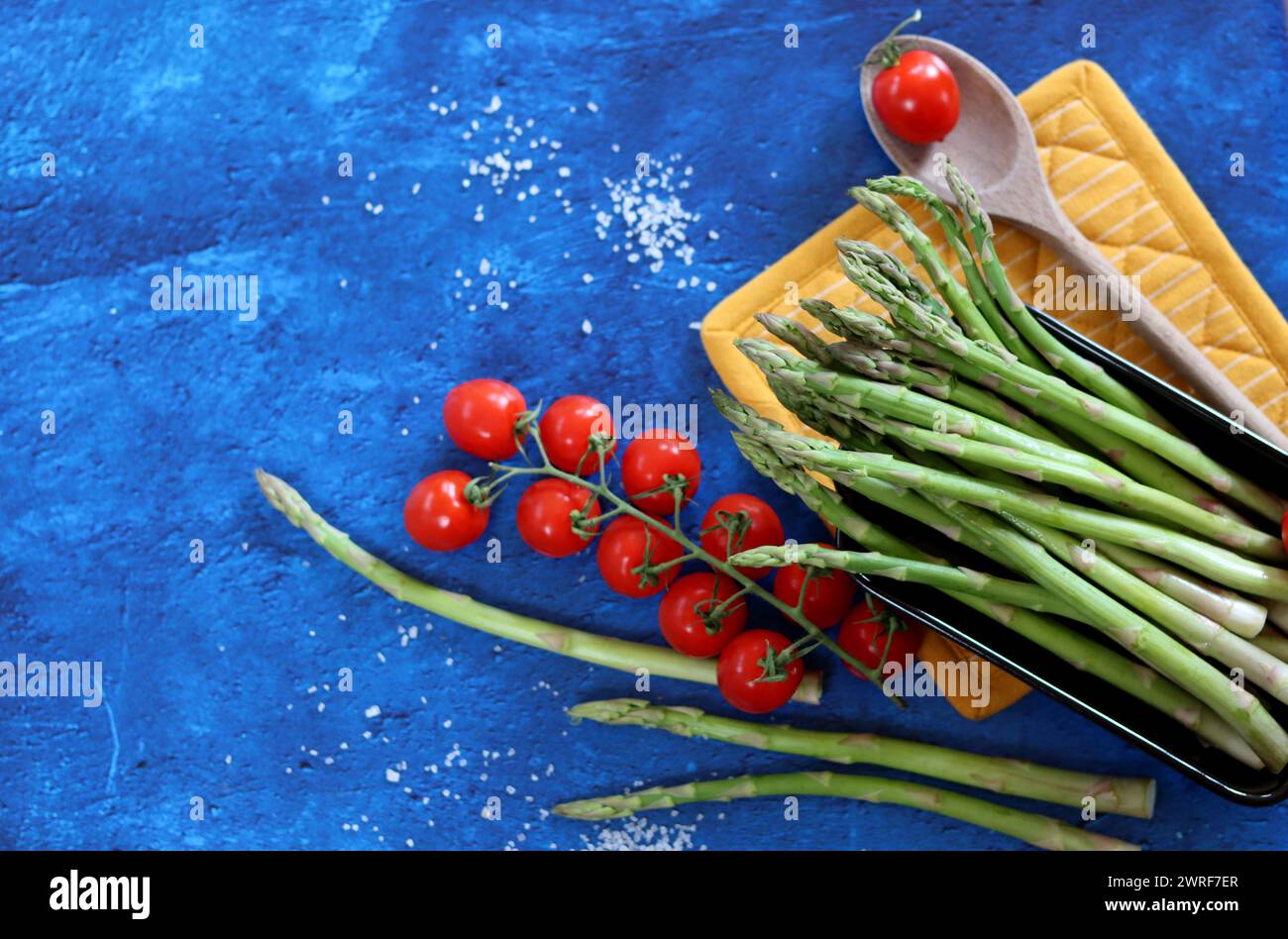 Frischer grüner Spargel und Kirschtomaten auf blau strukturiertem Hintergrund mit Kopierraum. Gesunde Zubereitung des Abendessens. Ausgewogenes Ernährungskonzept. Stockfoto