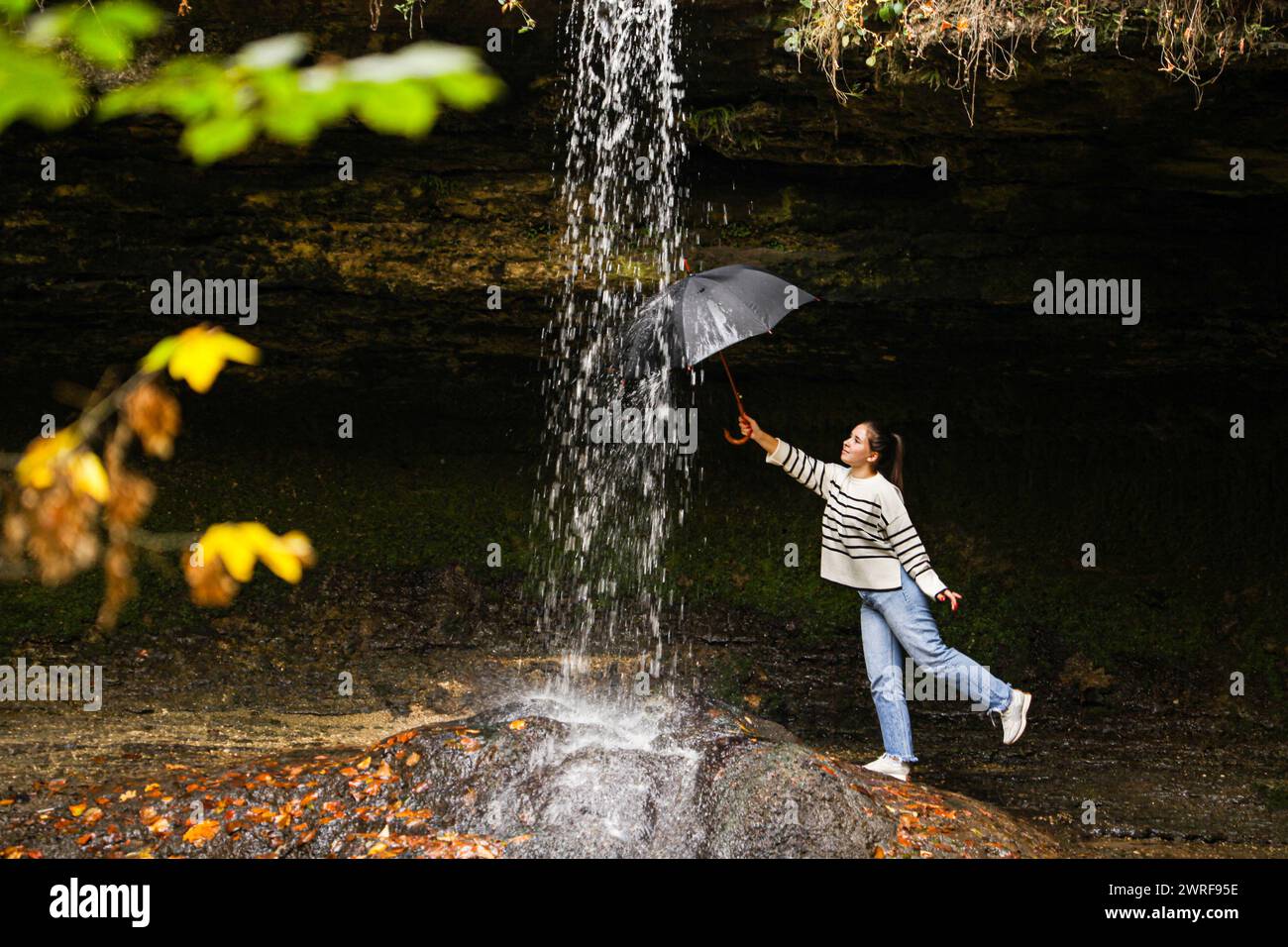 Eine Landschaft mit einem Wasserfall und einem Mädchen, das einen Schirm in einer ungewöhnlichen Position hält und ihn unter das Wasser stellen will, das aus dem Wasserfall fließt. A b Stockfoto