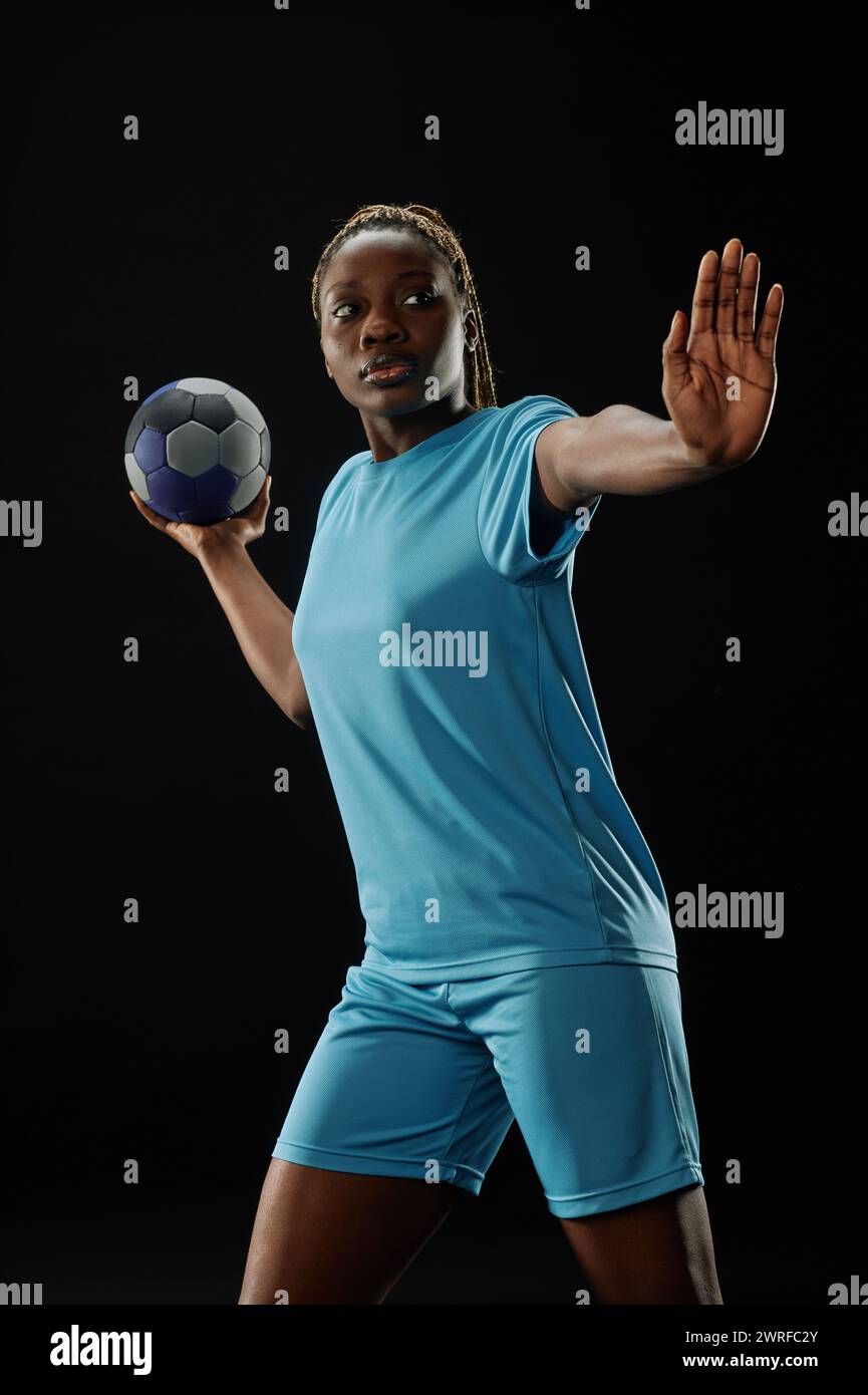 Dramatische Action-Aufnahme einer afroamerikanischen Fußballspielerin, die vor schwarzem Hintergrund mit intensivem Gesichtsausdruck den Ball vorbeiführt Stockfoto