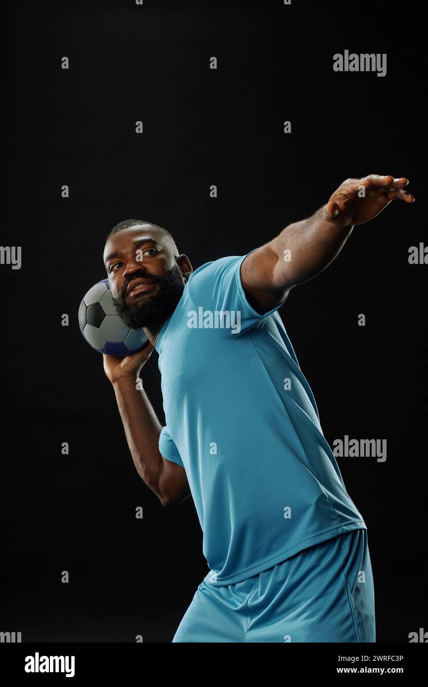 Vertikaler Action-Schuss eines afroamerikanischen Fußballspielers, der den Ball vor schwarzem Hintergrund mit intensivem Gesichtsausdruck übergeht Stockfoto