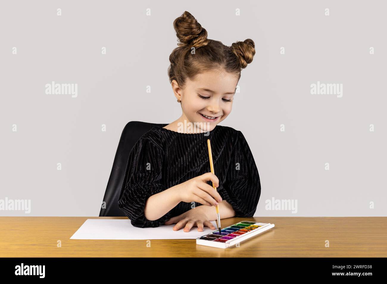 Ein kleines Mädchen sitzt an einem Tisch und lächelt, während sie mit Aquarellen malt. Sie ist umgeben von Kunstgegenständen und Schmuck und schafft ein wunderschönes Meisterwerk Stockfoto