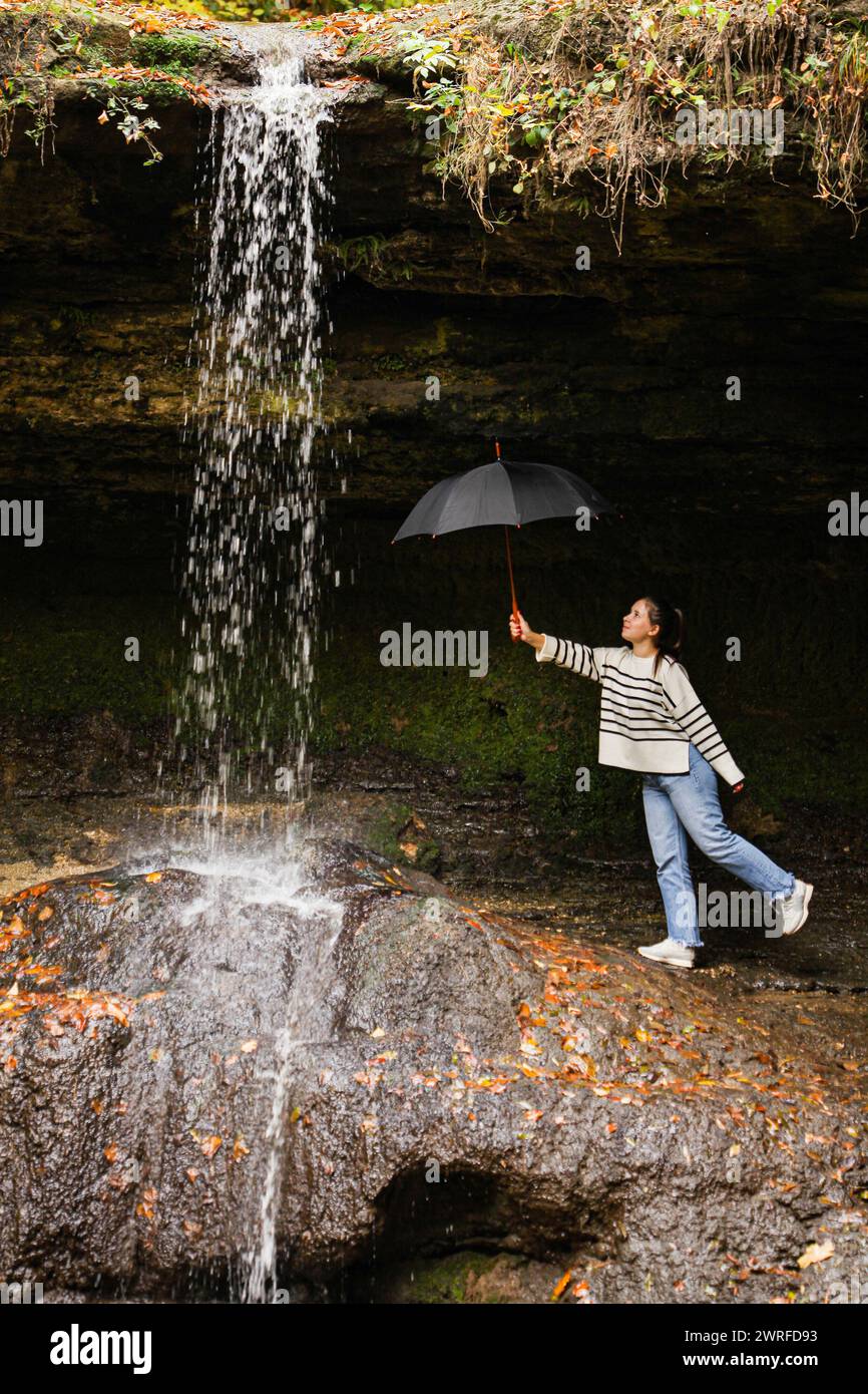 Ein glückliches junges Mädchen, das einen schwarzen Regenschirm unter den Wasserfällen hält, blickt zum Wasserfall und lächelt. Das Konzept, die Schönsten zu besuchen Stockfoto