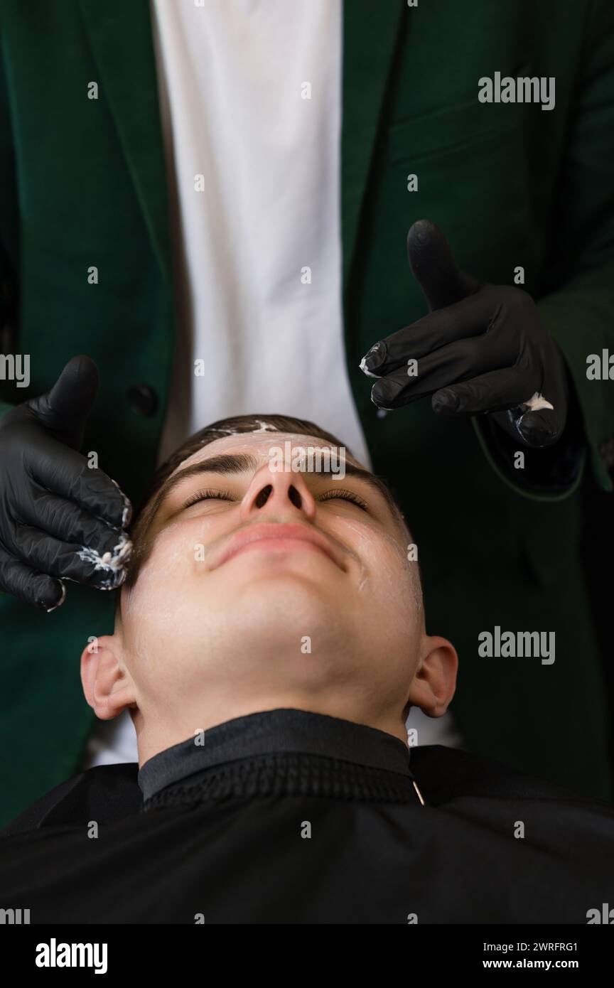 Ein Friseur in schwarzen Handschuhen trägt eine feuchtigkeitsspendende weiße Creme auf das Gesicht des Kunden auf. Eine Kosmetikerin trägt eine weiße Maske mit einem Vitaminkomplex auf die Haut von auf Stockfoto