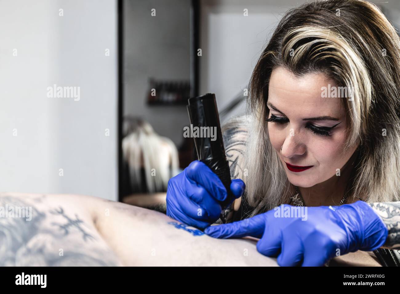 Horizontales Foto eine Nahaufnahme einer Tätowiererin, deren Blick auf die Haut fixiert ist, während ihre Hand die Tätowiermaschine mit fachkundiger Präzision über den Kunden führt Stockfoto