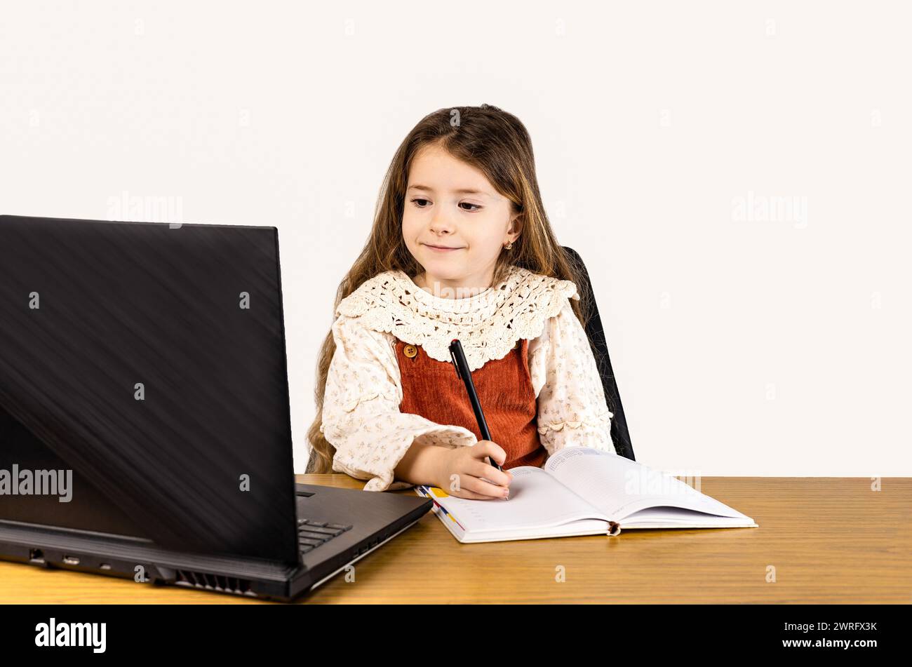 Ein junges Mädchen sitzt an einem Schreibtisch mit Laptop und Notebook, umgeben von Bürobedarf. Sie arbeitet an ihrem PC in einer Holzverkleidung Stockfoto