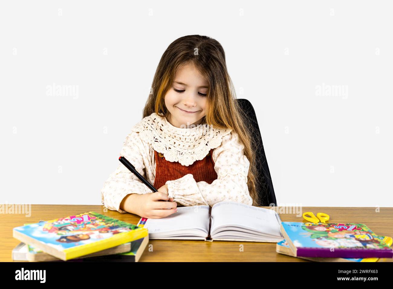 Ein glückliches Kleinkind sitzt an einem Schreibtisch, lächelt beim Schreiben in ein Notizbuch. Sie benutzt Bürobedarf und ein Schreibgerät. Teilen Sie ihre Freizeit t Stockfoto