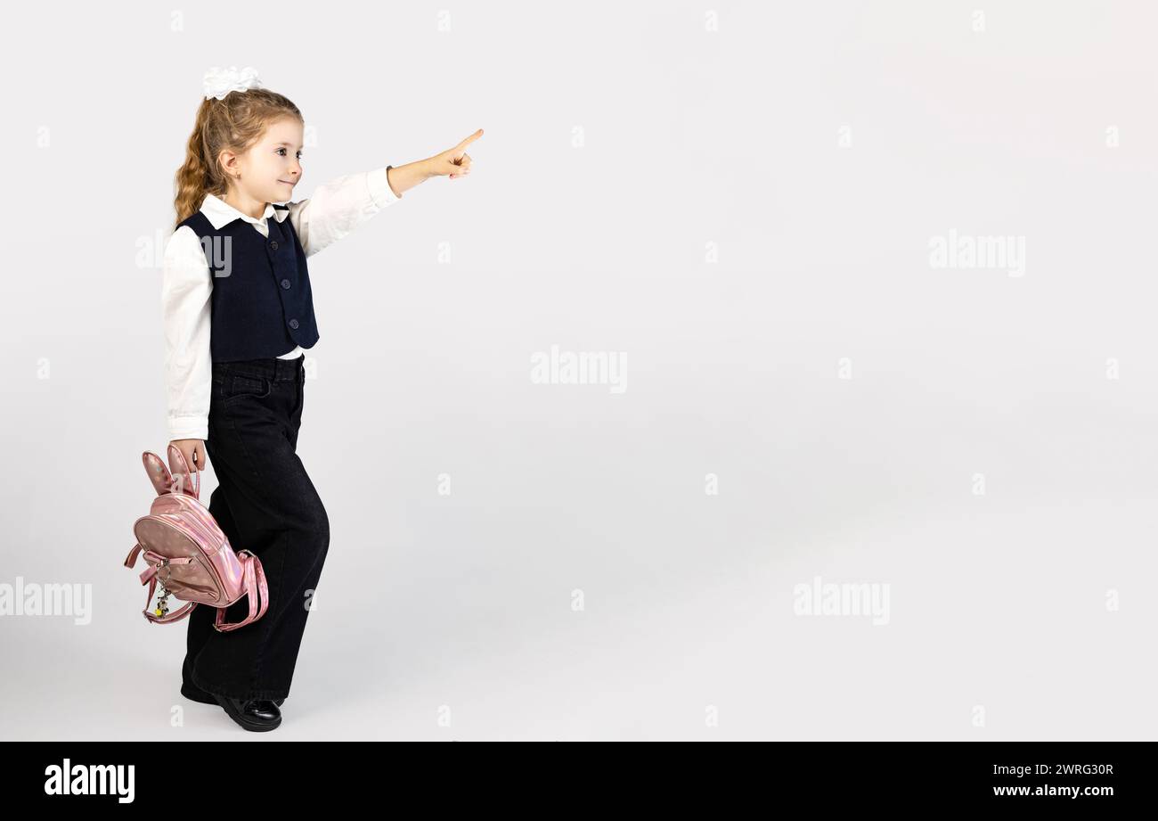 Ein junges Mädchen in Schuluniform zeigt glücklich mit dem Daumen auf etwas, während es einen rosa Rucksack hält. Sie steht mit einem Knie leicht Stockfoto
