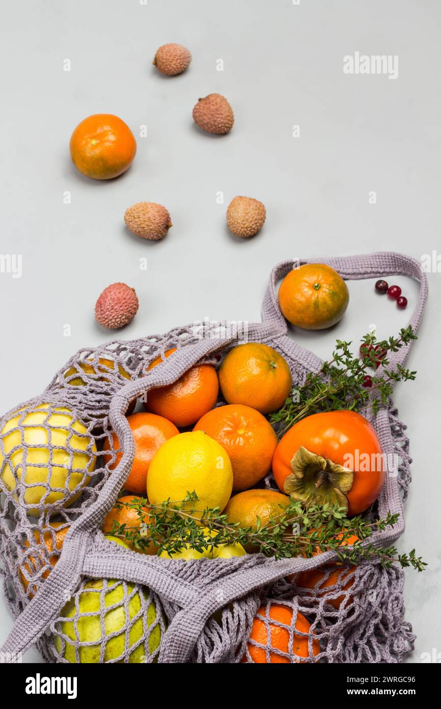 Persimmon, Äpfel und Zitrone im wiederverwendbaren Netzbeutel. Litschi, Preiselbeeren und Mandarinen auf dem Tisch. Grauer Hintergrund. Draufsicht Stockfoto