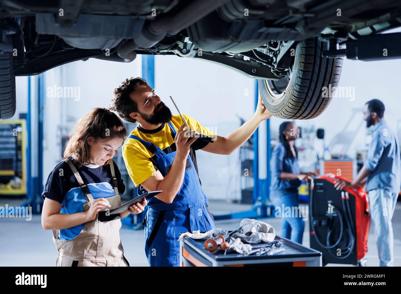 Techniker, die bei der Wartung eines defekten Fahrzeugs zusammenarbeiten und auf defekte Räder prüfen. Geschickte Werkstätten in der Autowerkstatt arbeiten zusammen an der Reparatur von Fahrzeugen und besprechen die besten Optionen Stockfoto