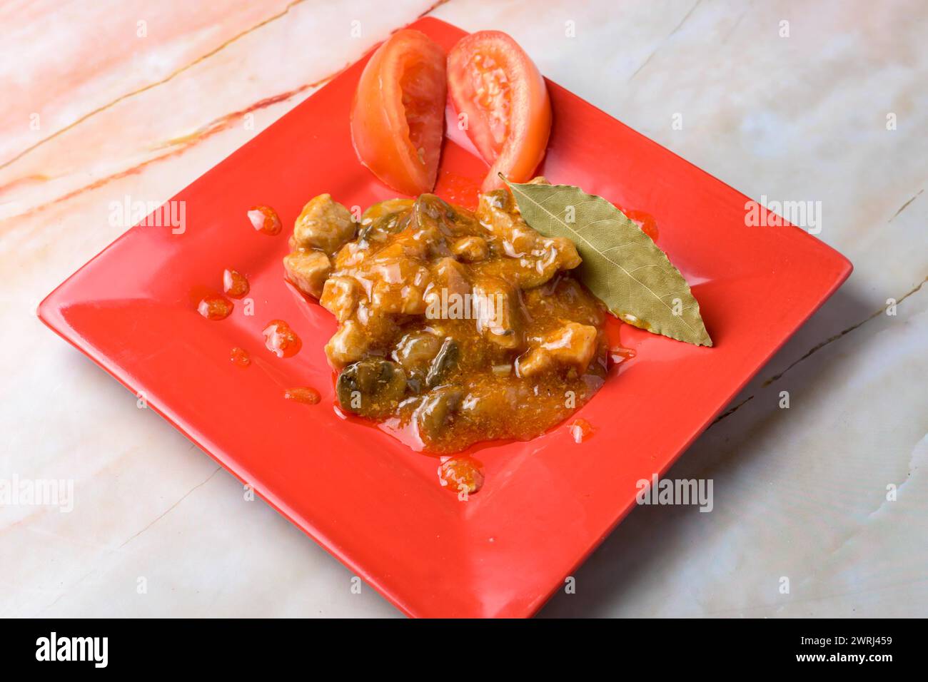 Schweineeintopf mit Lorbeerblatt und Tomatengarnitur auf rotem Teller, Marmorhintergrund, typisches Essen, typisch mediterrane mallorquinische Küche Stockfoto