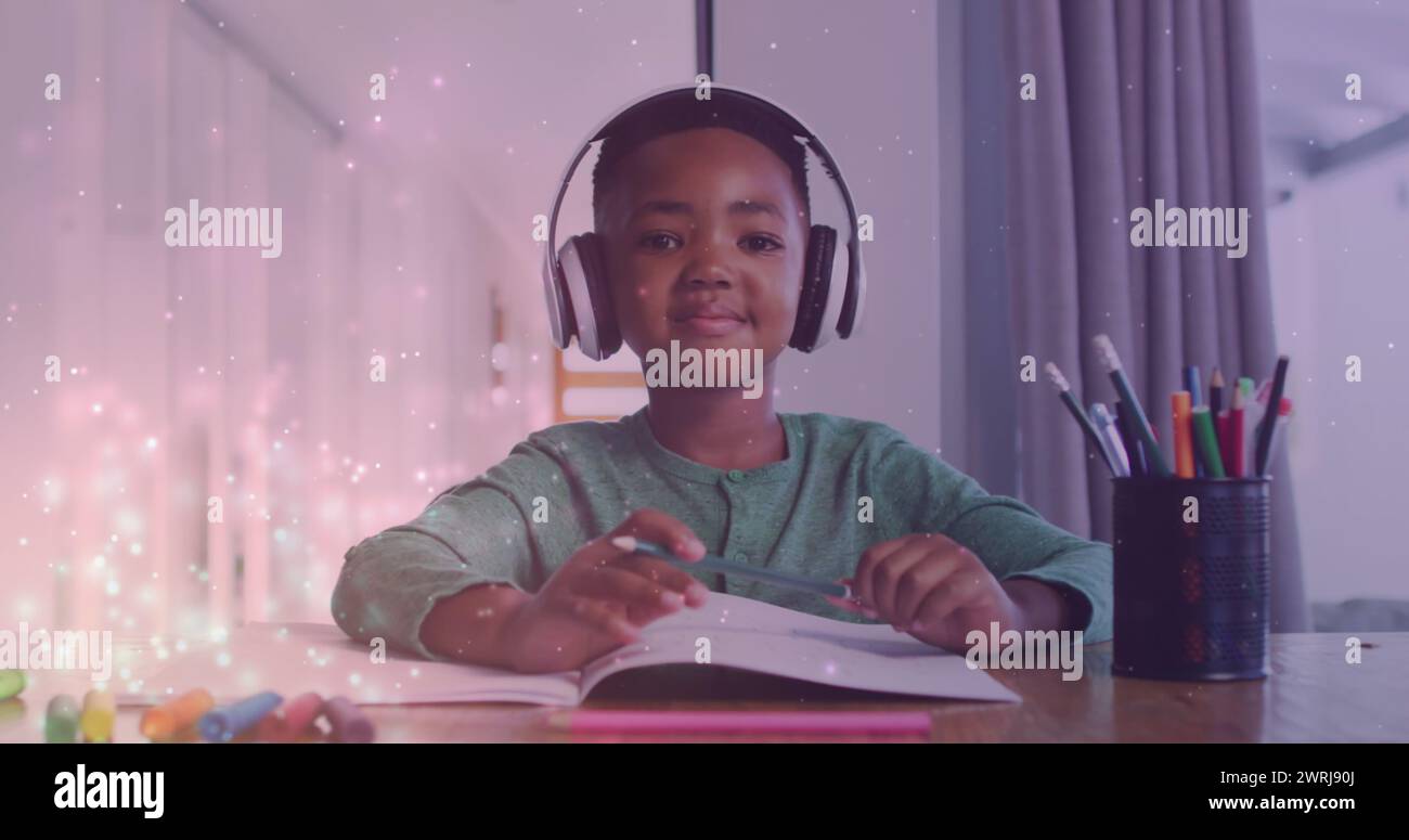 Bild von Lichtern über einem glücklichen afroamerikanischen Jungen, der Kopfhörer trägt und Hausaufgaben macht Stockfoto