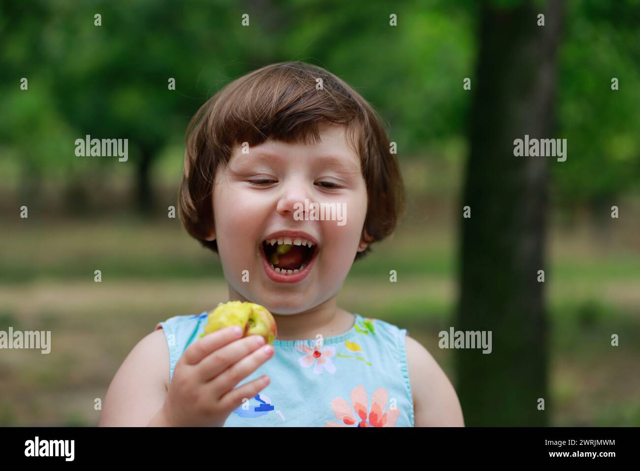 Niedliches kleines Mädchen, das unter Obstbäumen spielt, in Sommerkleid gekleidet. Verschwommene grüne Bäume im Hintergrund. Unter Obstbäumen spielen und Obst essen Stockfoto