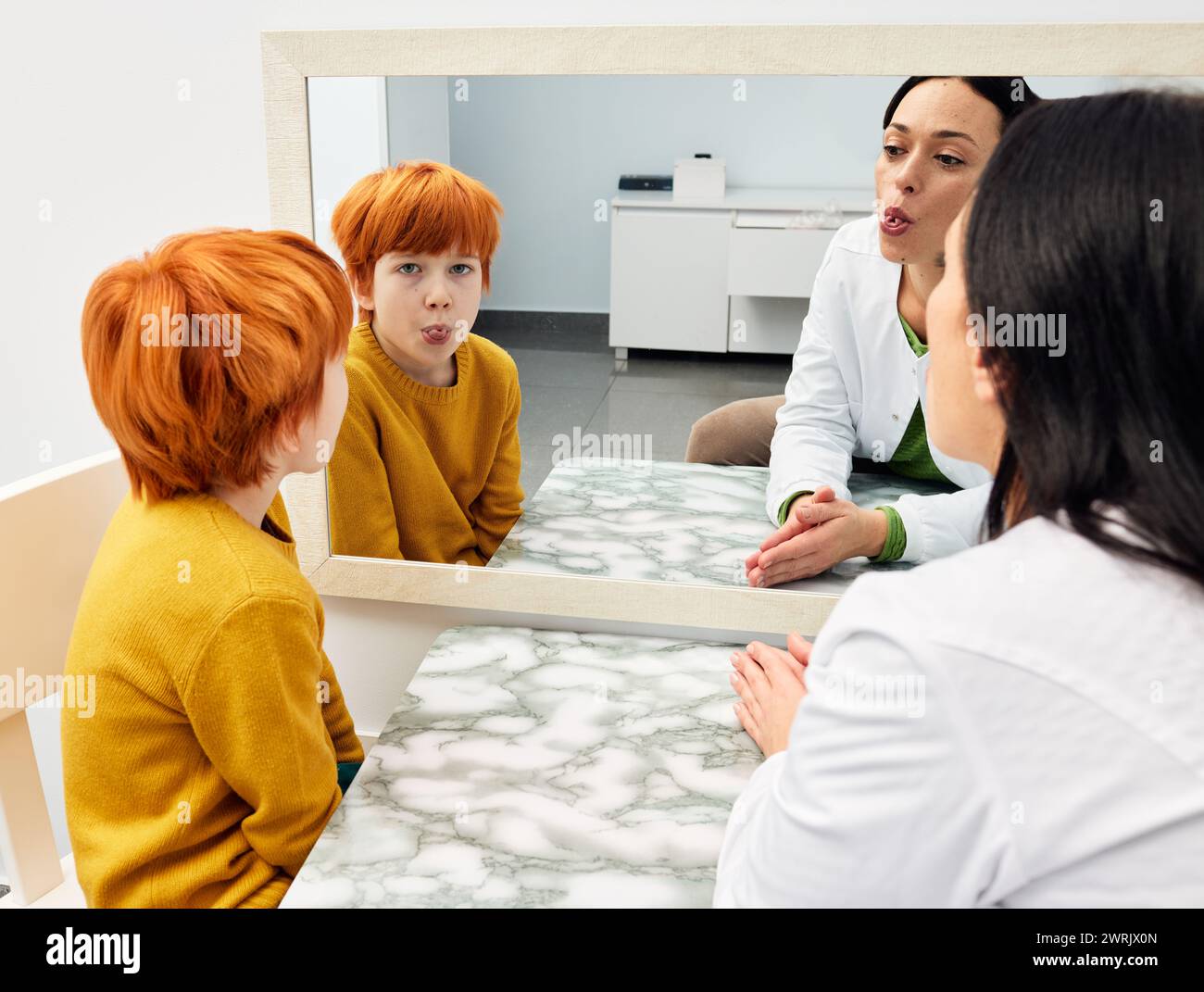 Sprachtraining. Junge, der Zungenübungen mit seinem Logopäden in der Nähe eines großen Spiegels für die richtige Aussprache macht Stockfoto