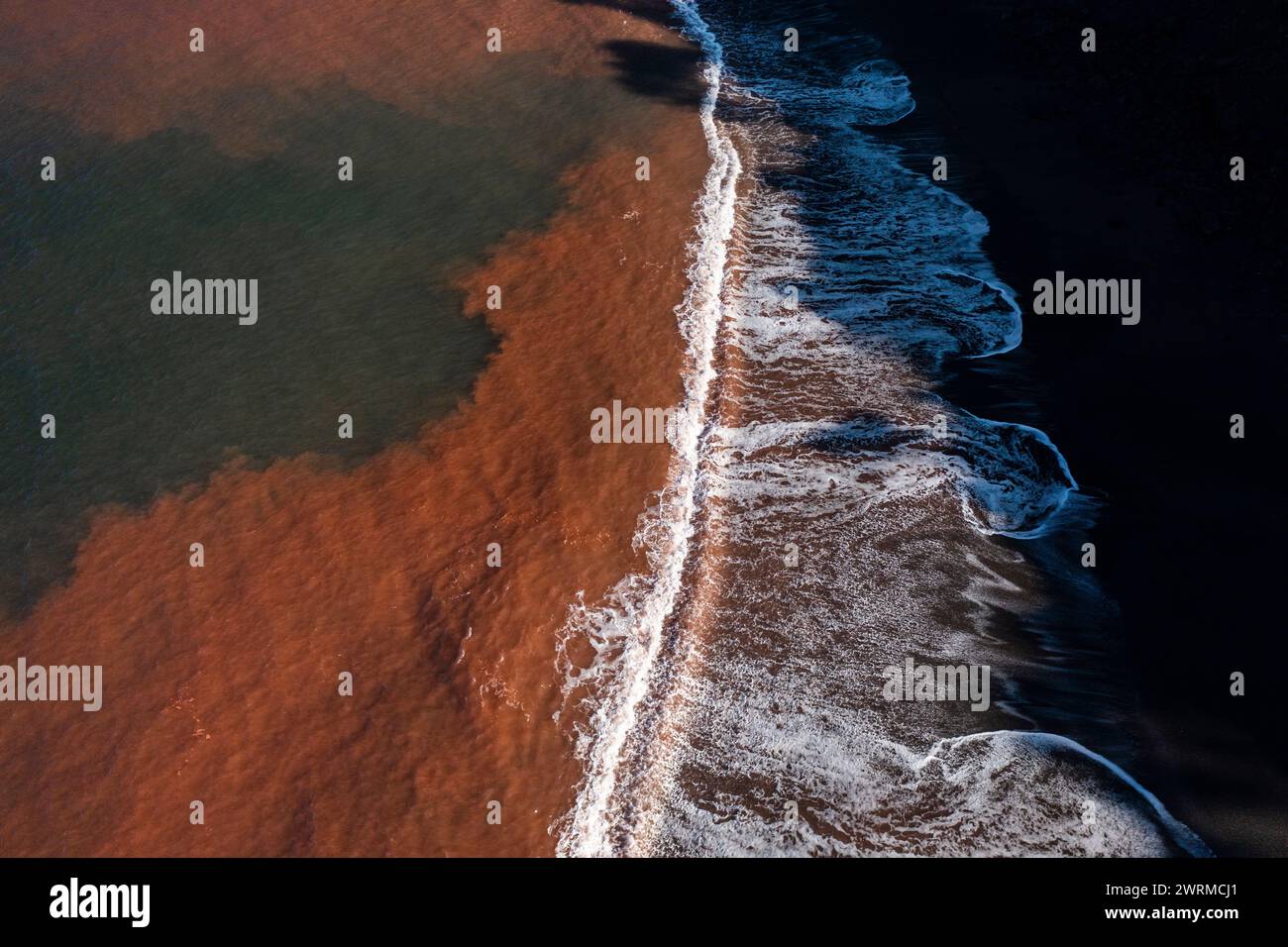 Luftaufnahme des starken Kontrasts zwischen dem rötlichen, mit Eisenoxid befleckten Wasser und dem unberührten weißen Schaum der Wellen am Strand Llumeres in Asturien, an Stockfoto