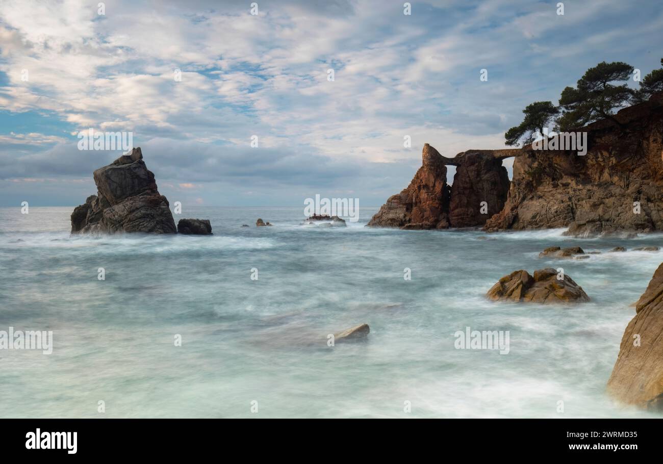 Langzeitaufnahmen, die das ruhige Wasser und die felsige Landschaft von Cala dels Frares an der Costa Brava mit bewölktem Himmel über Ihnen einfangen. Stockfoto