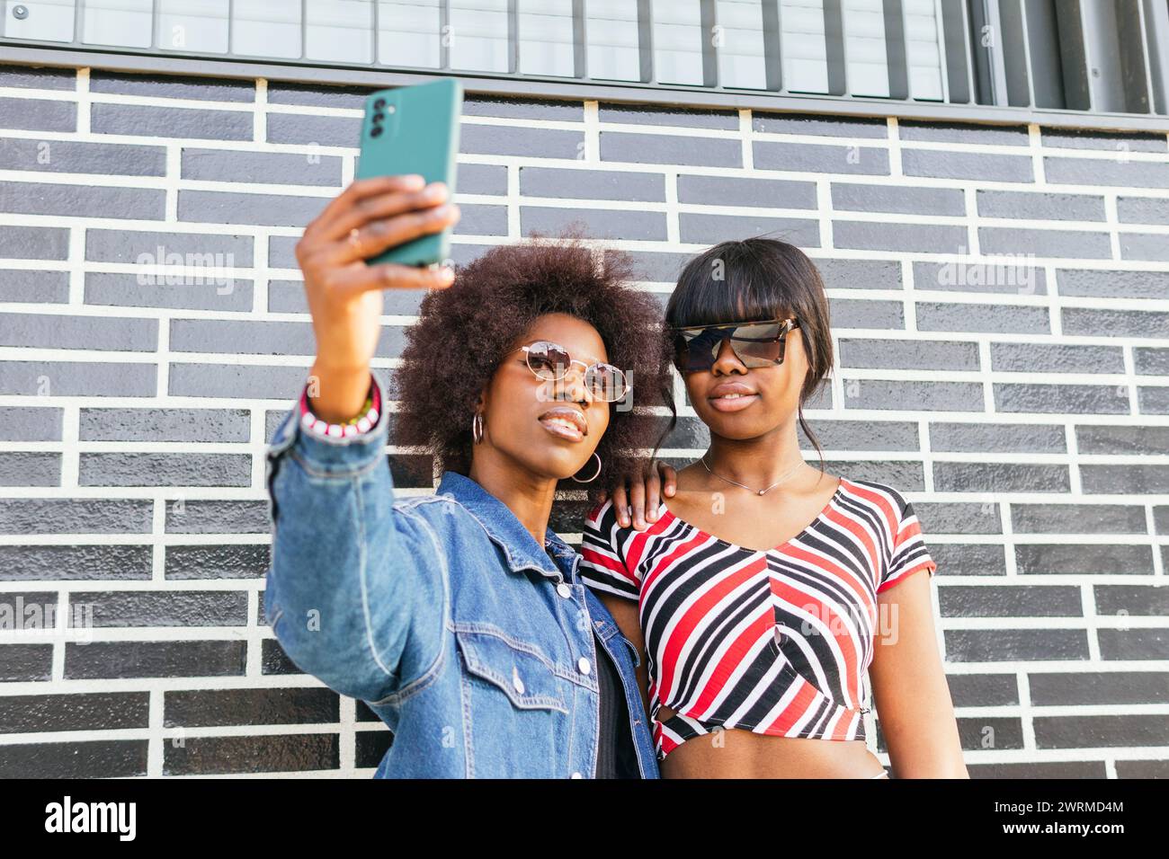 Zwei Schwestern machen ein Selfie, um einen Moment ihres pulsierenden urbanen Lebens zusammen vor einer markanten Ziegelmauer festzuhalten Stockfoto