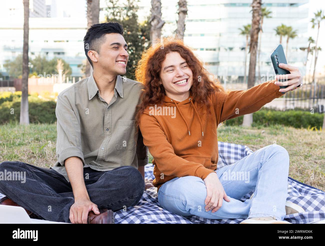 Zwei glückliche Studenten, eine junge Frau mit lockigen roten Haaren und ein junger Mann lateinischer Herkunft, halten einen freudigen Moment mit einem Smartphone-Selfie fest, während sie sitzen Stockfoto